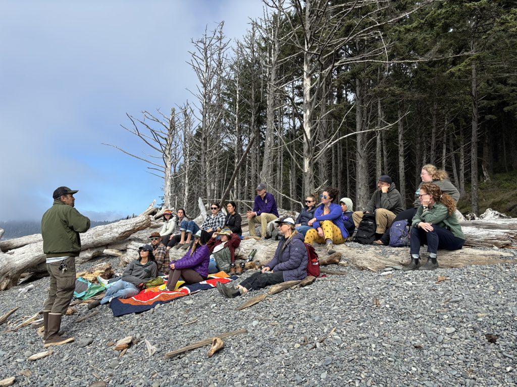 Dr. Steve Fradkin, standing, addressed a group of seated fellows on a rocky beach, with forest beyond.