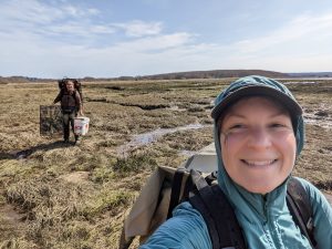 A selfie of Alex in the field on a coastal marsh with Alexa in the background carrying a bucket and shrimp traps