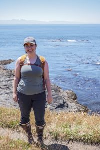 Staci standing on a ledge on Lopez Island. She is standing in front of the ocean with bull kelp at the surface.