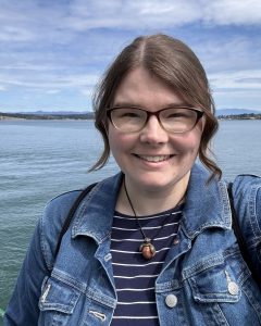New WSG science communications fellow Emily Bjornsgard smiles for a photo, wearing brown rimmed glasses, a denim jacket, and a striped shirt in front of the calm waters of Puget Sound.