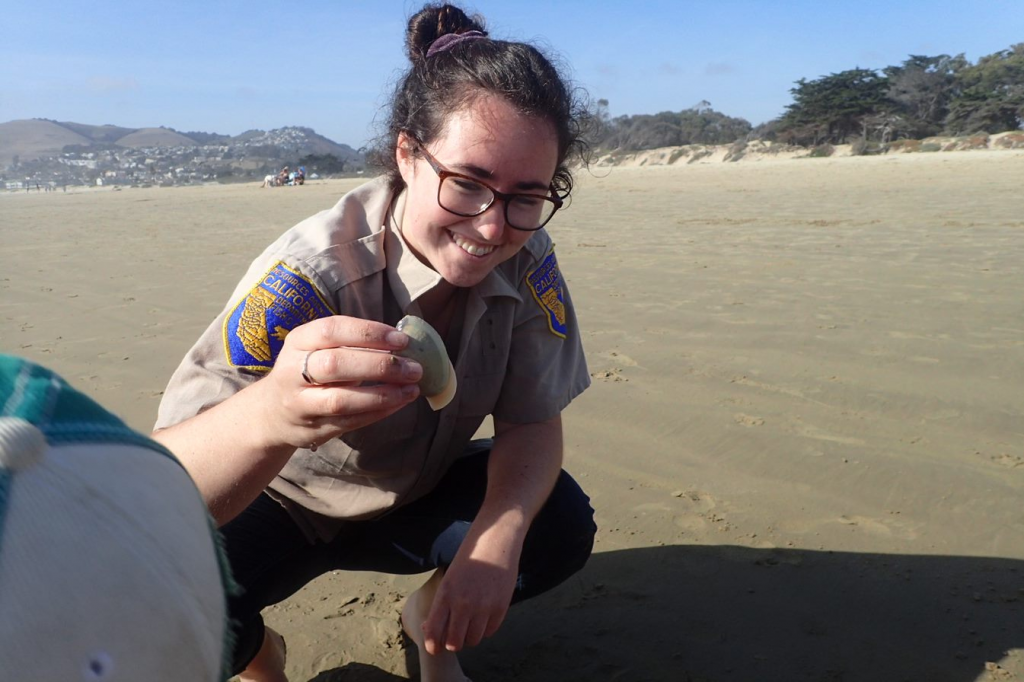 Hannah is crouched down on a wide, sandy beach and smiling at a three-inch Pismo clam that she is holding in her hand. The Pismo clam’s foot is out of the bottom of its shell. The foot looks a bit like a tongue.