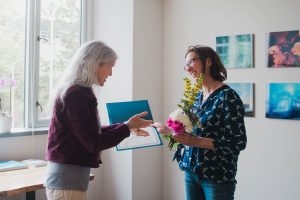 Maya Tolstoy presents a certificate to Kate LItle, who is holding a bouquet of flowers.