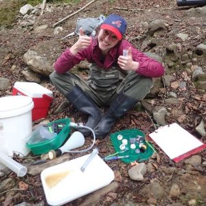 Lisa in waders, sitting amidst various tubes and buckets alongside a stream, grinning and giving two thumbs up.