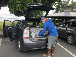 A photo shows a person standing in front of an open tailgate of a Prius. In the trunk is a water filtration set up, with a vacuum pump run by the car's battery, and filtering eDNA water samples.