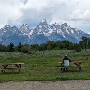 Lisa sitting at a picnic table, facing the Teton Mountains in the distance which span the width of the image