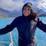 Mariah Ricci on a boat deck with blue water, mountains and clouds behind.