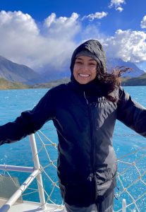 Mariah Ricci on a boat deck with blue water, mountains and clouds behind.