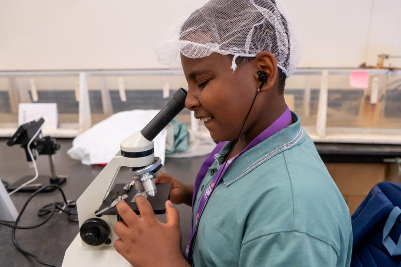 A student wearing a hairnet smiles looking into a microscope.